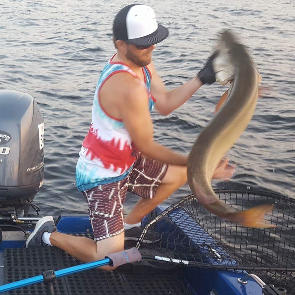 Guy pulling a muskie out of a fishing net while kneeling on a fishing boat